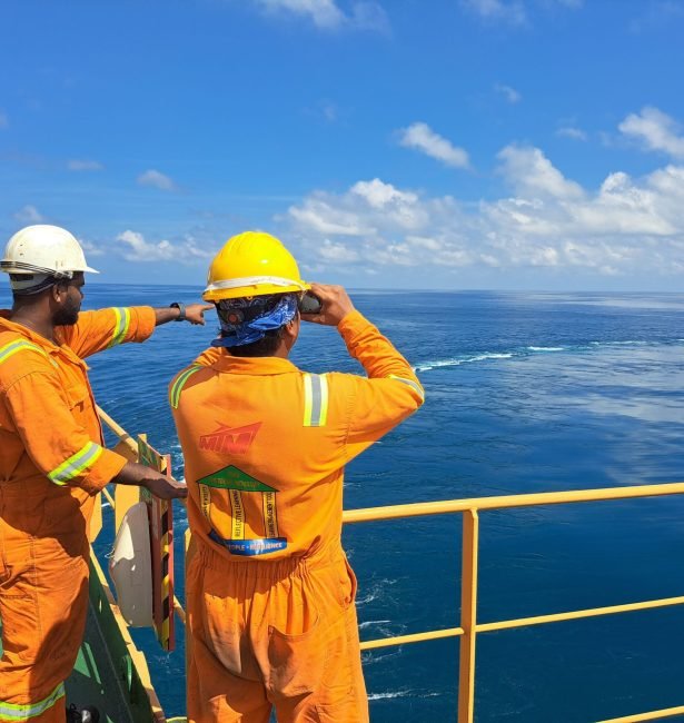 Two workers on an oil rig deck wearing safety gear inspect the ocean horizon under a clear sky.